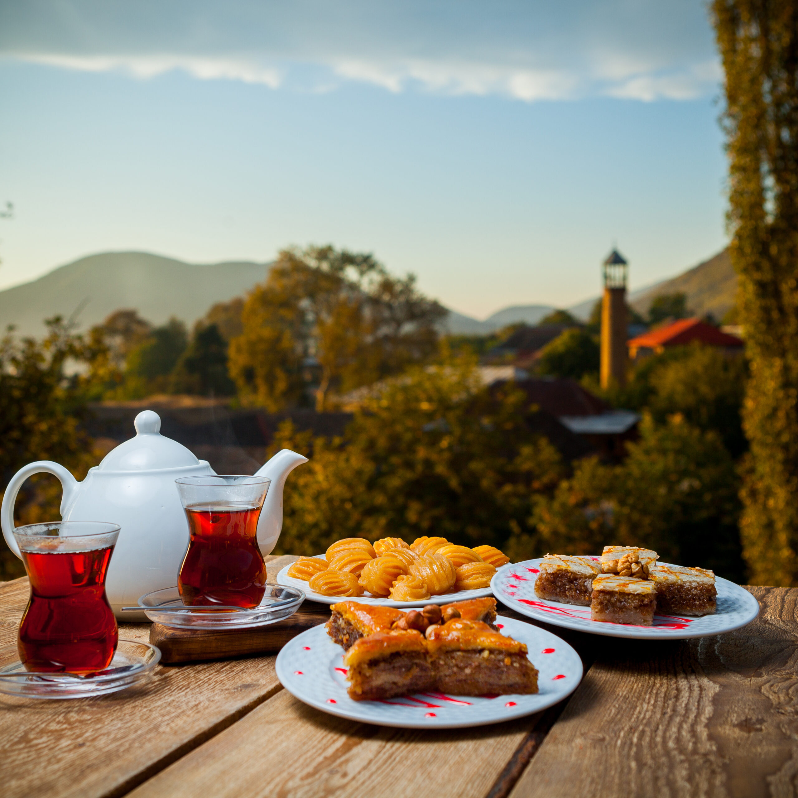 Some turkish desserts with glasses of tea and teapot on a table with village on background, side view.